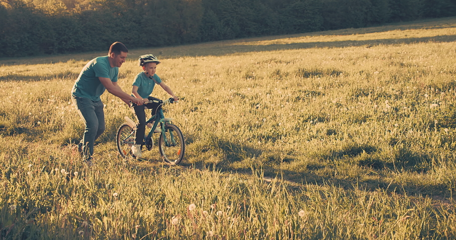 Father teaching son to ride bicycle. Happy cute boy in helmet learn to riding a bike in park on green meadow in summer day at sunset time. Family weekend. 4K video Slow motion