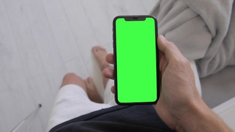 Man hand holding iPhone 11 with blank green screen chroma key and browsing social networks in the morning routine. POV footage, first person view, indoor. 2020 - Malibu, USA