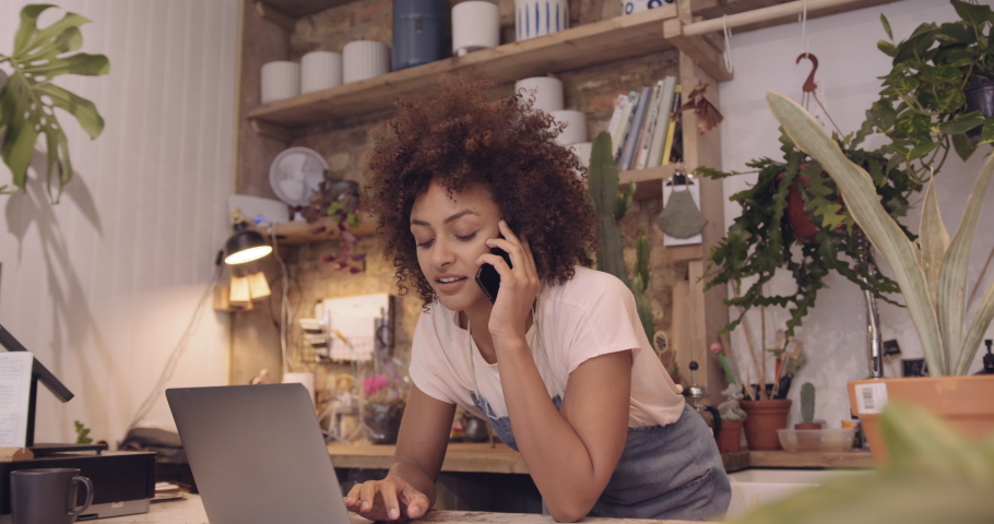 Young adult Afro American female small business owner working on laptop in store and speaking to client on phone Royalty-Free Stock Footage #1056918833