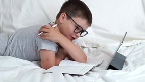 schoolboy in glasses using digital tablet device. Child making notes in copybook doing school homework lying on sofa. Online learning, distance lesson, education with online tutor at home quarantine.