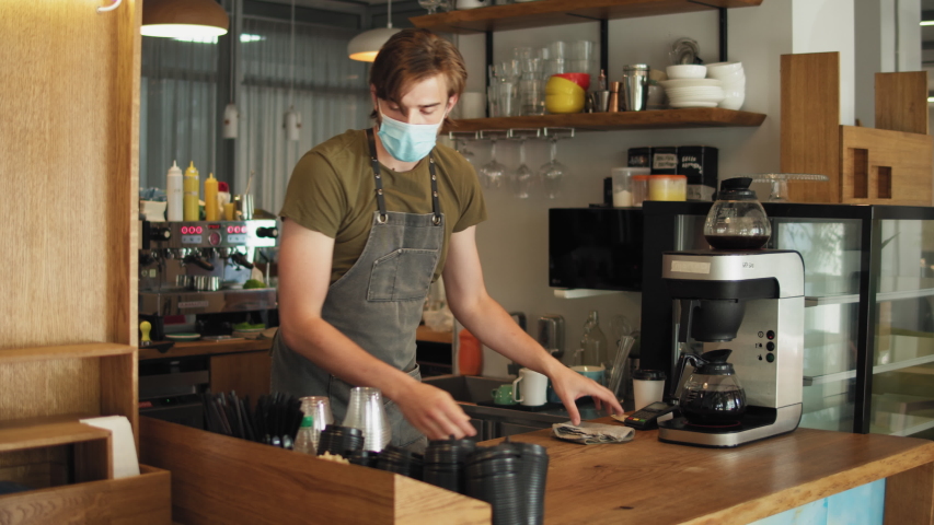 young barista man in mask and apron cleaning and preparing bar for visitors working in coffee shop, sanitizer desinfection during coronavirus covid 19 pandemic, reopen business, post covid Royalty-Free Stock Footage #1056932570