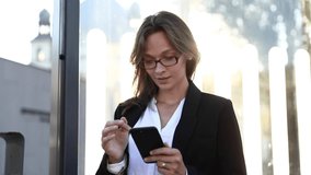 Happy business lady in eyeglasses and office outfit chatting with friends in social networks while waiting for transport. Beautiful woman using smartphone for online conversation.