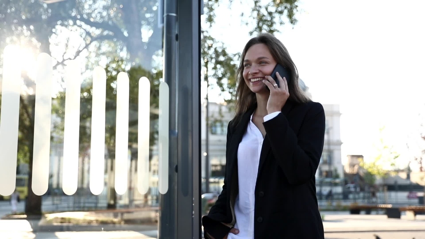 Happy woman in formal clothing chatting with friend on smartphone outdoors. Smiling girl with brown hair walking on street near business center and talking on mobile during free time.