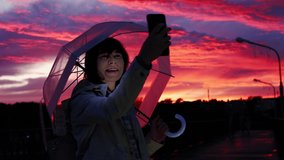 Young caucasian woman with a transparent umbrella makes selfie using a smartphone at dusk on a background of pink-blue clouds.