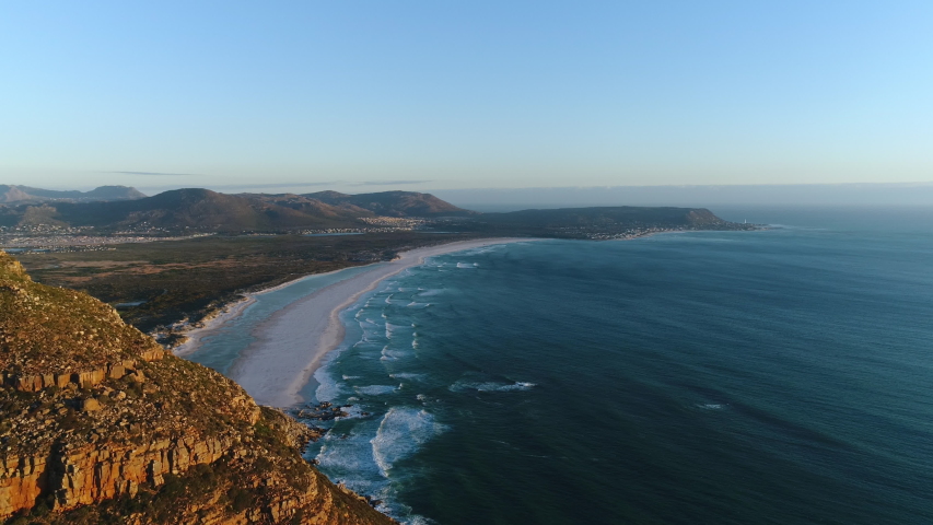 Aerial view of Noordhoek's large white beach and flying back to reveal the scenic mountain pass of Chapman's peak. People taking a sunset drive along one of the most iconic coastal roads in the world. Royalty-Free Stock Footage #1056942203