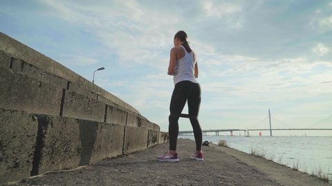 Fit young woman doing squat exercises outdoors at the sunset. Slow motion of athletic girl training with elastic band in urban city background. Active and sport concept Stockvideó