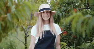Portrait of female horticulturist in hat and apron holding wicker basket full of ripe peaches. Happy woman with blond hair standing at garden, smiling and looking at camera