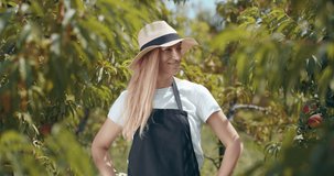Portrait of smiling beautiful woman in summer hat posing on camera with crossed arms at peach garden. Female farmer in protective apron enjoying season work outdoors.