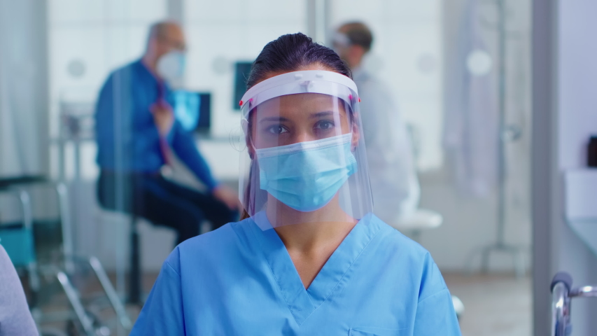 Medical assistant with visor and face mask against coronavirus looking at camera in hospital waiting area. Doctor consulting senior man in examination room. | Shutterstock HD Video #1056958289