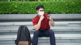 close up young asian man sitting at the park and wear face mask while using video call form smartphone to talking with friends and family for social distance new normal and healthy lifestyle concept