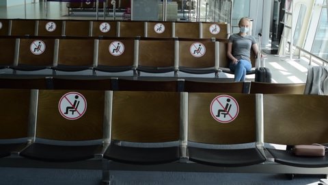 Woman tourist wearing medical protection mask sits in airport terminal with social distancing marker seat informing individuals that certain seats are closed during COVID-19.