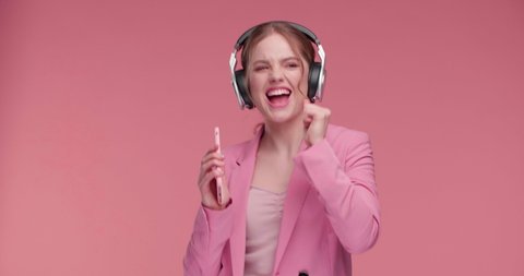 Amazing Girl is sincerely enjoying listening to Music in modern Headphones. Red-haired Girl starting listening to Music, using new Music App on Smartphone. Colorful pink Frame on Studio. Cute Girl.