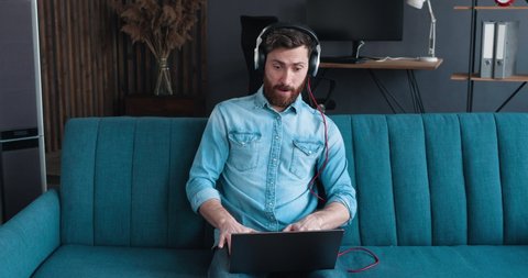 Male Gamer wearing Headphones, playing a Video Game on his Laptop PC in Living Room. Joyful Bearded Man is playing Emotional on Sofa at Home. Playing Games in Quarantine. Gamers Lifestyle. Indoor.