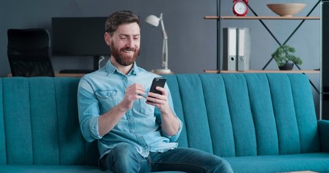 Satisfied Man with Beard in casual Shirt is sitting in Loft Living Room and using his Smartphone. Handsome Man feels Happy at Home, surfing Internet, smiling. Home Quarantine. Standing Indoor.
