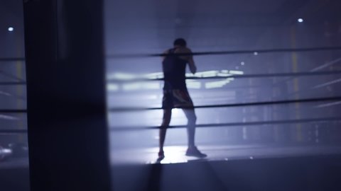 Kickboxer or Muay thai fighter training in low light gym . Silhouette on dark background . Boxer training in ring . Cinematic slow motion shot of a professional young man practicing in boxing ring