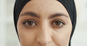 Close up of face of young Arabian woman in black hijab looking straight in camera at home. White background. Portrait. Muslim beautiful female in traditional Arabic headscarf.