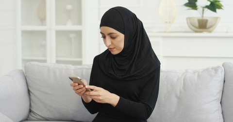 Cheerful young Arabian beautiful woman in black hijab sitting on couch and texting message on smartphone at home. Pretty smiled female muslim in headscarf tapping and scrolling on mobile phone.
