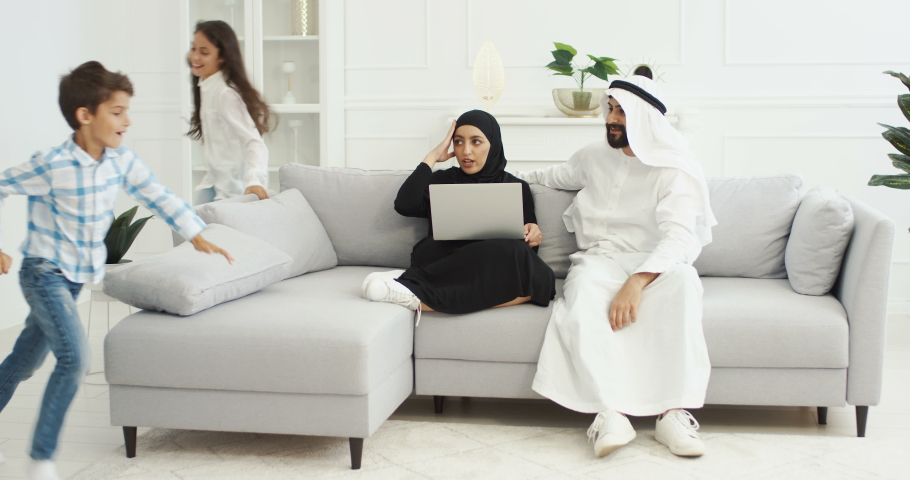 Cheerful Arabic children running in cozy living room. Happy little kids, sister and brother playing around sofa. Muslims mother and father sitting on couch with laptop computer at home and smiling.