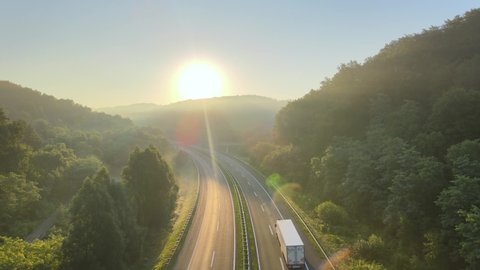 Isolated white truck drive alone on a empty highway road delivering supplies during an emergency pandemic virus crisis, aerial footage sunset over wild nature