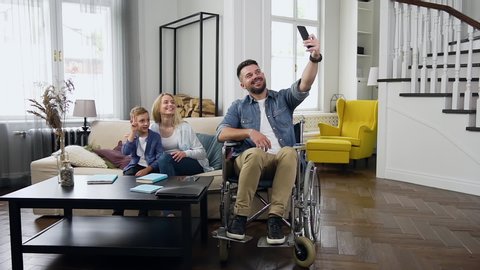 Handsome smiling bearded disabled 30-aged man taking photos on his smartphone together with his splendid contented wife and 8-aged son in beautifully designeted apartment