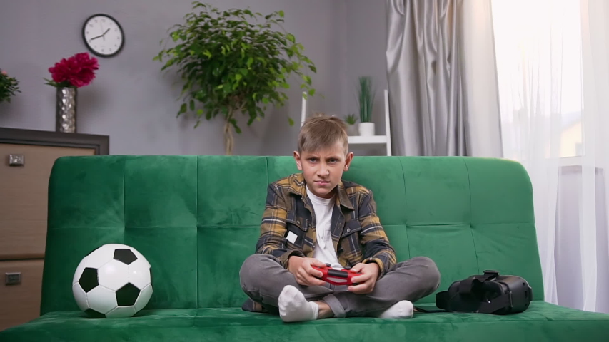 Good-looking confident serious 10-aged boy spending his leisure at video gaming with gamepad while sitting on soft green couch in contemporary living-room