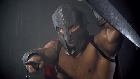 Front motion of fearless muscular spartan hiding from rain under shield. Ancient warrior shirtless in iron helmet and red cloak pointing spear in bad rainy weather, bright light. Concept of warrior.