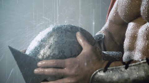 Close up motion of raindrops bouncing on iron helmet in hands of muscular spartan wearing red cloak and armour. Close up of wet sexy handsome caucasian man posing in bad weather. Concept of warrior.