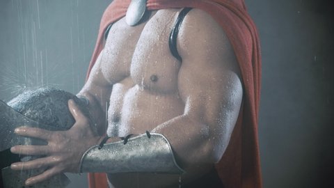 Motion of unrecognizable muscular spartan wearing red cloak, holding iron helmet, raindrops bouncing. Close up of wet sexy handsome caucasian man posing in bad weather. Concept of warrior.