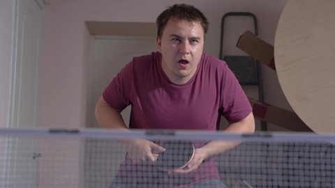Young caucasian male play ping-pong at home or club, indoors. Man wearing casual t-shirt hits the tennis ball and sends it somewhere, after covers his mouth with hand in horror, making big eyes