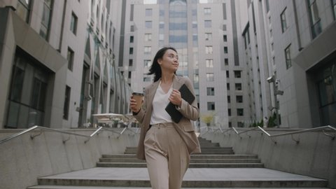 Successful young beautiful asian woman with tablet and coffee cup walking between business buildings and smiling with joy. Attractive business girl. Career people.