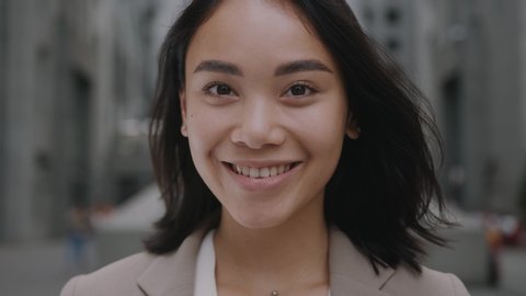 Face portrait of attractive joyful young asian mixed race woman in business formal suit smiling and posing near modern buildings outside. Businesswoman. Ethnicity.