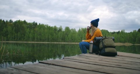 A woman with a hat and a backpack sits on a wooden pier of a forest lake in the mountains and drinks hot drinks tea or coffee from a thermos.