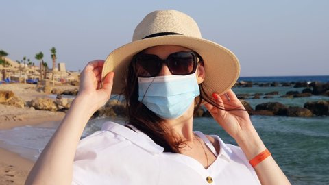 portrait of woman in protective mask, sunglasses and sun hat, against sea background, beach with palm trees. sunny warm day. Travelling after Covid-19. end of self-isolation.