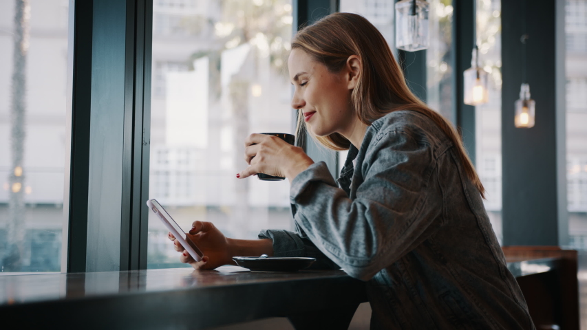Woman sitting at a coffee shop with mobile phone drinking coffee and looking away. Caucasian female relaxing at a cafe.
 | Shutterstock HD Video #1056988910