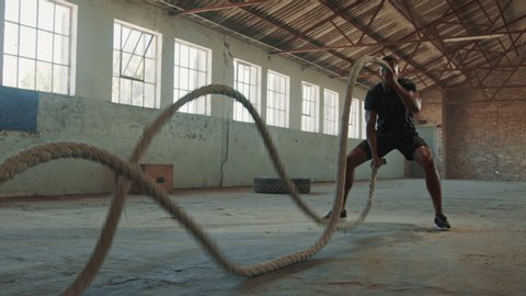 Fitness man exercising with battle rope abandoned warehouse. Tough man working out in cross training gym made inside old factory.
