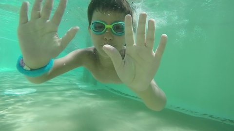The boy in swimming goggles is swimming at the bottom of the pool and waving hands underwater