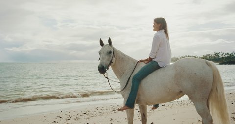 Woman riding beautiful white horse down the beach in slow motion, relaxing coastline