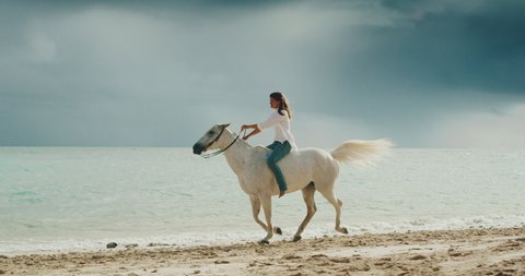 Woman horseback riding, galloping down sandy coastline on beautiful white horse in cinematic slow motion