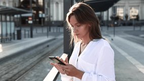 Serious business woman in white shirt working on modern smartphone while waiting for bus. Beautiful dark-haired girl holding digital gadget on street.
