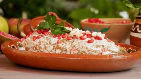 Chile en Nogada Mexican dish in traditional clay plate with pomegranate seeds and ingredients on wooden table, panning motion on food
