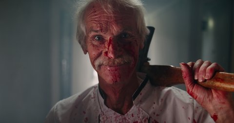 Mustached senior caucasian psycopath creepily staring at camera and grinning, wearing butcher's apron covered in blood and carrying bloodstained axe - halloween concept 4k portrait
