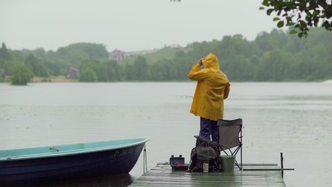 Tracking tilt up back view shot of bearded old fisherman in yellow raincoat fishing in lake standing on wooden pier in rain
