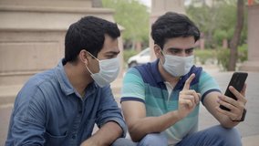 Two young male colleagues wearing face protective mask sitting together outdoors and using mobile phone or smartphone. Close friends discussing  looking at the cellphone amid Corona virus epidemic