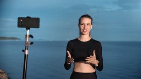 Female online coach demonstrating stretching training in live broadcasting use smartphone. Smiling blogger woman shooting video of workout on beach explaining exercise. 4k Dragon RED camera