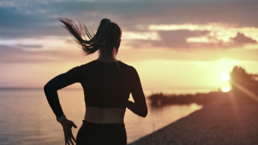 Back view sportswoman running at seashore with dramatic sky and bright sunshine slow motion. Silhouette of female jogger during outdoor activity on beach. Medium shot on RED camera