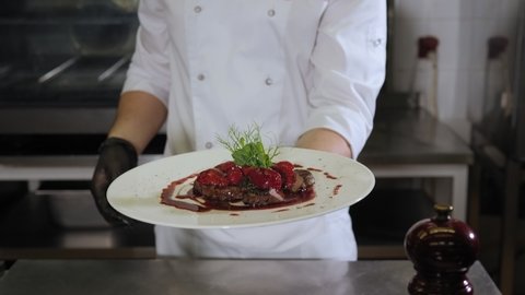 A uniformed chef holds a plate of beef steak and strawberry sauce in the restaurant's kitchen. Serving meat dishes with berry sauce in the restaurant. Slow motion.