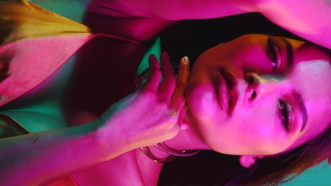 Portrait shot of young flirting cute Asian woman playfully posing for camera on the pink and blue neon lights - vertical video in slow motion