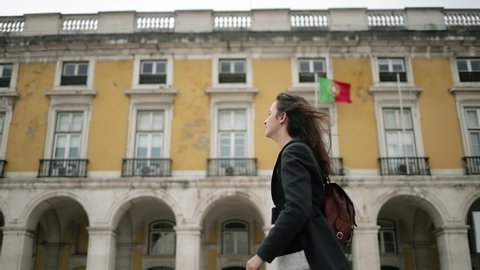 Pretty young female tourist in overcoat and backpack walking past state building with Portuguese flag. Side view, low angle. Travel in Portugal concept