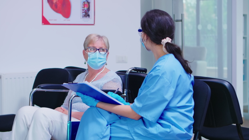 Nurse explaining diagnosis to disabled senior woman in wheelchair. Assistant wearing face mask against infection with coronavirus in hospital waiting area. | Shutterstock HD Video #1057021721