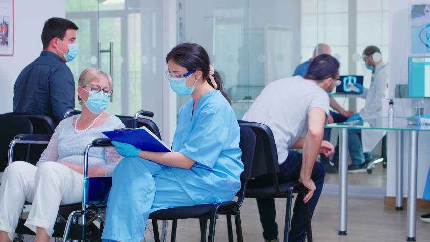 Disabled senior woman in wheelchair talking with nurse that wears protection mask against infection with coronavirus. Patient and medical staff in waiting area. Doctor in examination room. | Shutterstock HD Video #1057021799